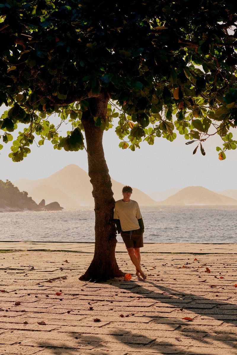 Model Francesco Ruggiero leans against a tree trunk on a paved seaside promenade overlooking coastal waters and distant hills.