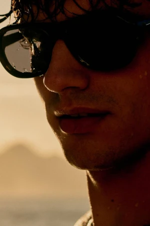 Close-up portrait of a man wearing dark sunglasses against a bright coastal sunset sky background for Zara Man beachwear.