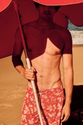 Shirtless man standing on a sandy beach under a red sun umbrella wearing red floral print swim trunks.