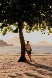 Model Francesco Ruggiero leans against a tree trunk on a paved seaside promenade overlooking coastal waters and distant hills.