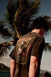 Man from behind wearing a washed dark graphic t-shirt in front of a palm tree and ocean background.
