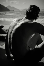 Monochrome shot of model Francesco Ruggiero seated shirtless on a sandy beach looking out toward the ocean waves.