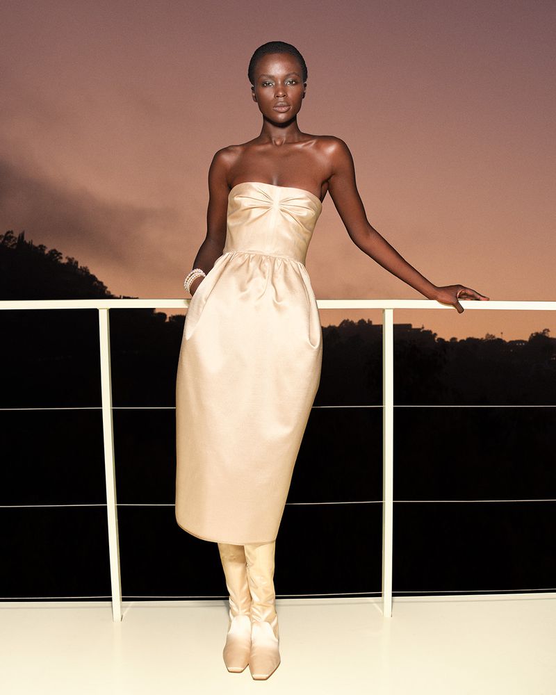 Model Awar Odhiang posing on a rooftop balcony wearing a strapless cream dress against a sunset sky.