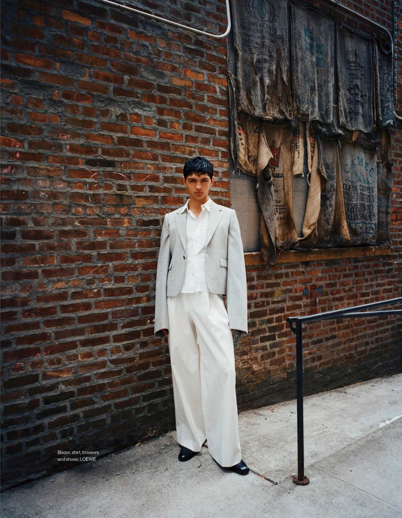 Fashion shot of a young man in a light grey jacket and white pants standing against a brick wall with faded posters in an urban environment.