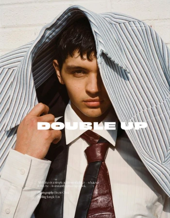 Portrait of a young adult man in a white button-down shirt and tie, with a striped coat draped over his shoulders against a white brick wall.