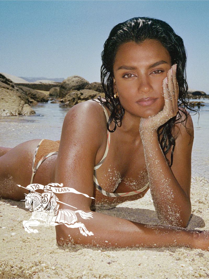 Simone Ashley lying on a sandy beach wearing a check bikini for the Burberry x Hunza G campaign.