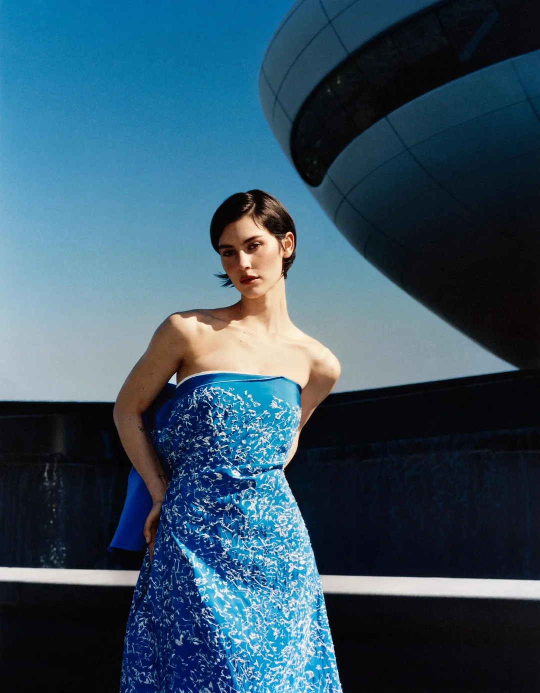 Gracie Abrams wearing a blue strapless dress posing outdoors against a modern architectural structure and clear blue sky.