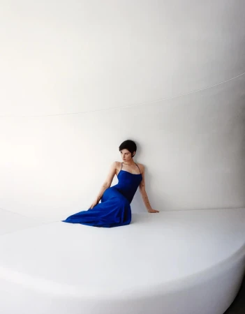 Gracie Abrams seated in a royal blue floor-length gown against a curving minimalist white architectural wall.