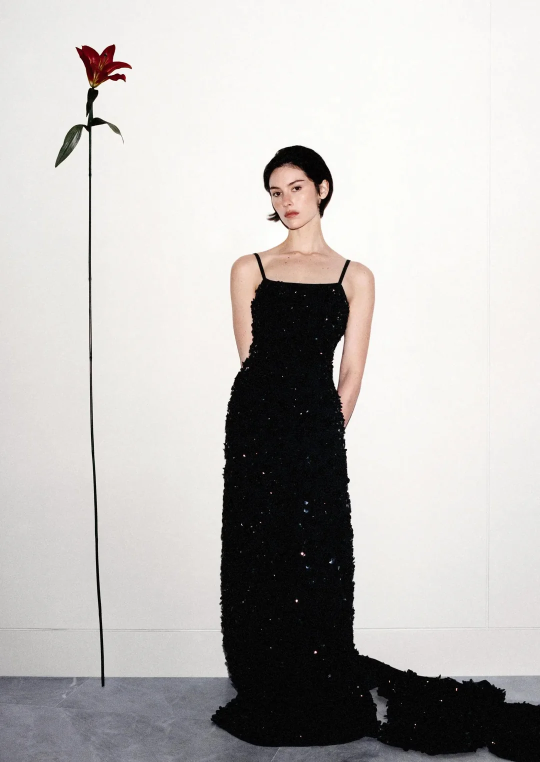 Gracie Abrams stands in a black sequined floor-length dress beside an oversized red flower against a white studio wall.