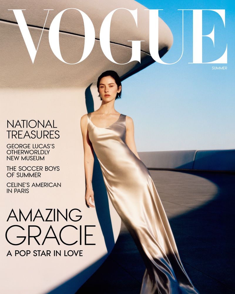 Gracie Abrams wearing a flowing satin slip dress leaning against a curved modern architectural structure