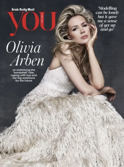 Model Olivia Arben posing in a strapless textured feather gown against a grey studio backdrop for a magazine cover.