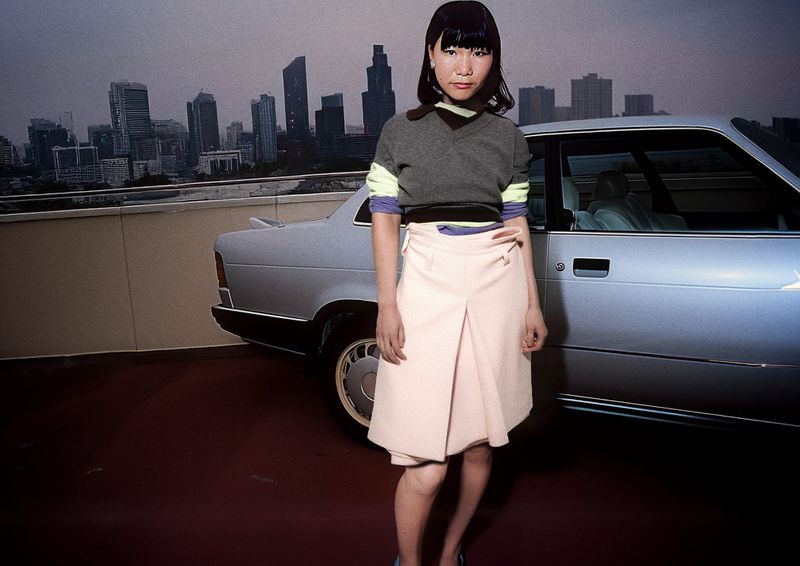 Adult woman poses in front of a silver car and cityscape backdrop, wearing a gray top, pink skirt, and blue heels.