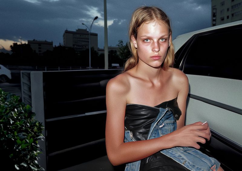 Portrait of a young blonde woman in a black leather top and denim jacket, standing next to a white car against an overcast sky.