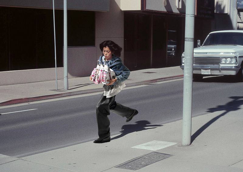 Woman runs across a city street holding a large, patterned handbag. A white truck is parked on the side of the road.