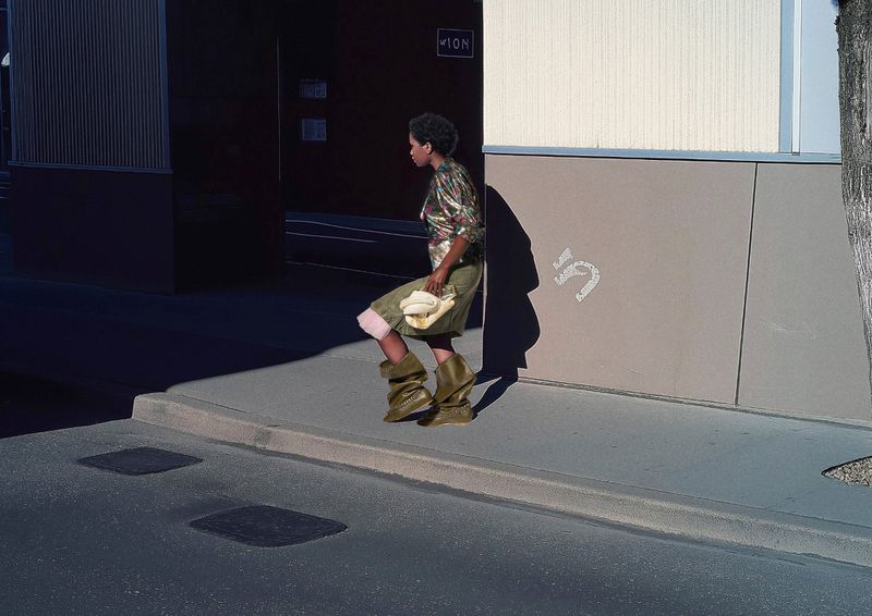 Woman in olive-green avant-garde boots walks near a building with a white handbag. The photo captures an urban fashion statement.