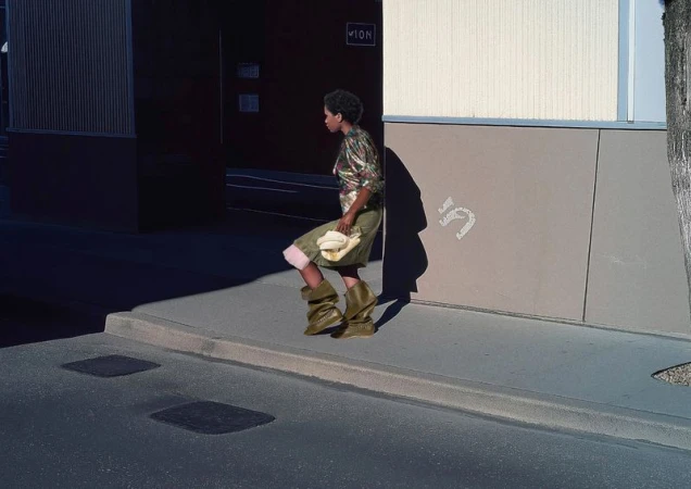 Woman in olive-green avant-garde boots walks near a building with a white handbag. The photo captures an urban fashion statement.