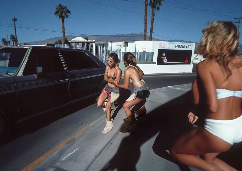 Three young women in swimsuits and short skirts stand on a roadside next to a black car in a desert environment with palm trees.