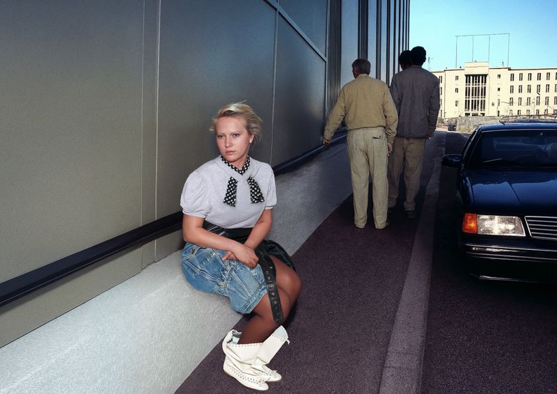 Blonde woman with polka dot blouse sitting outside on a ledge near a modern building with people and a car in the background.