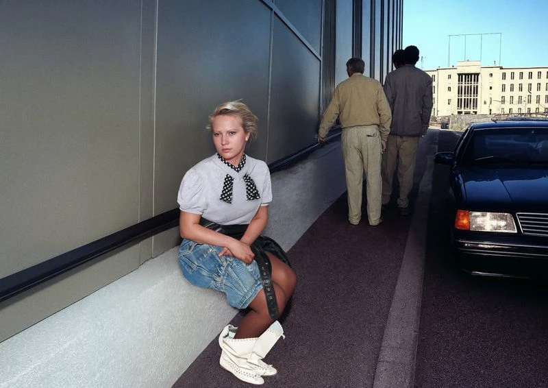 Blonde woman with polka dot blouse sitting outside on a ledge near a modern building with people and a car in the background.