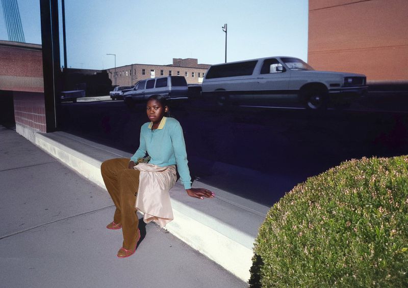 An African-American woman poses on a sidewalk in tan pants and blue top, with blurred cars in the background, and a green bush to the side.