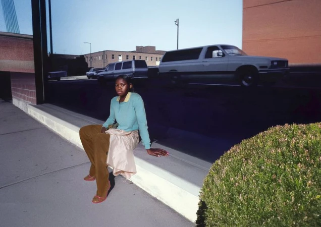 An African-American woman poses on a sidewalk in tan pants and blue top, with blurred cars in the background, and a green bush to the side.
