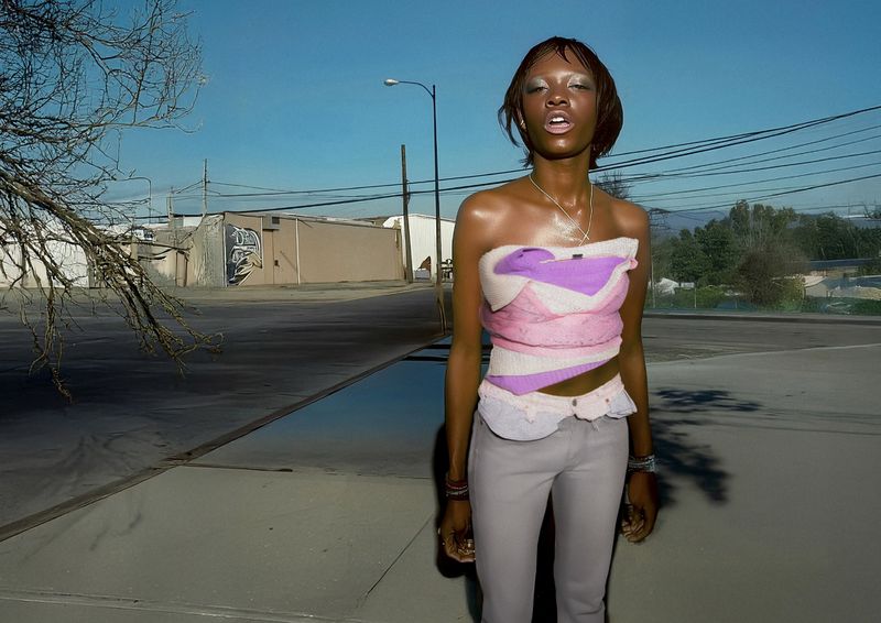 A slender African American woman wearing a colorful top and grey pants stands on a sidewalk, with industrial buildings in the background under a bright sky.