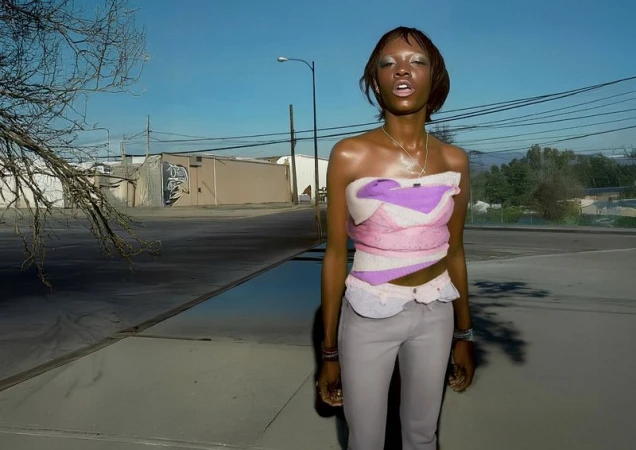 A slender African American woman wearing a colorful top and grey pants stands on a sidewalk, with industrial buildings in the background under a bright sky.