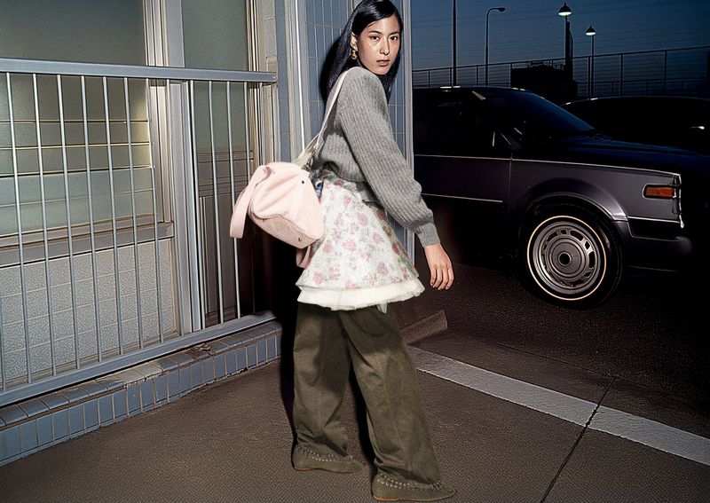 Asian woman in gray sweater, floral skirt, and platform shoes poses with a pink bag in an urban setting with cars in the background.