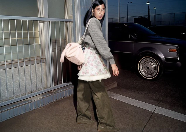 Asian woman in gray sweater, floral skirt, and platform shoes poses with a pink bag in an urban setting with cars in the background.