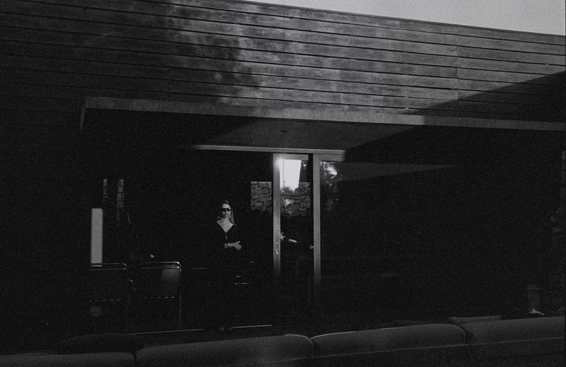 Woman in dark sunglasses standing in deep shadow under the overhang of a modern wood-paneled house in black and white.