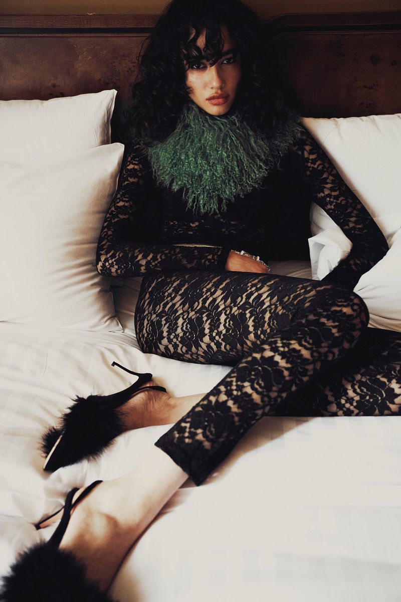 Fashion photo of an adult female model in lace bodysuit, tights, and heels, reclining on a white bed with a dark headboard.