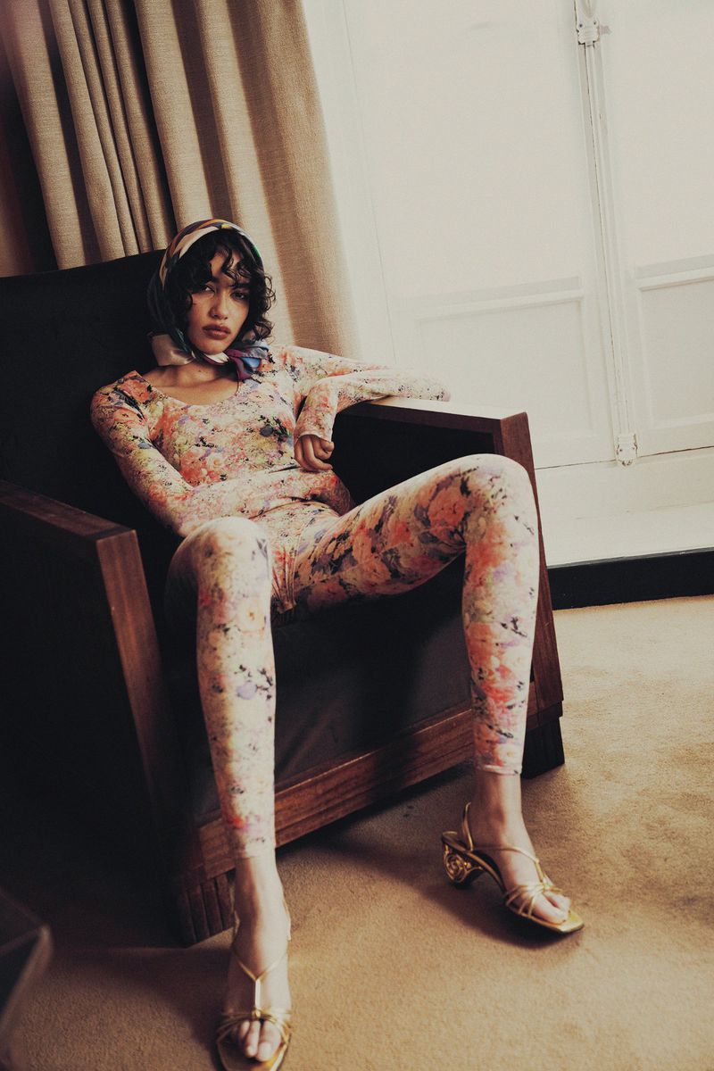 Woman in a floral bodysuit and headscarf sits in a dark wood chair indoors, with a window and light curtains in the background.