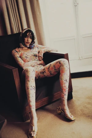 Woman in a floral bodysuit and headscarf sits in a dark wood chair indoors, with a window and light curtains in the background.