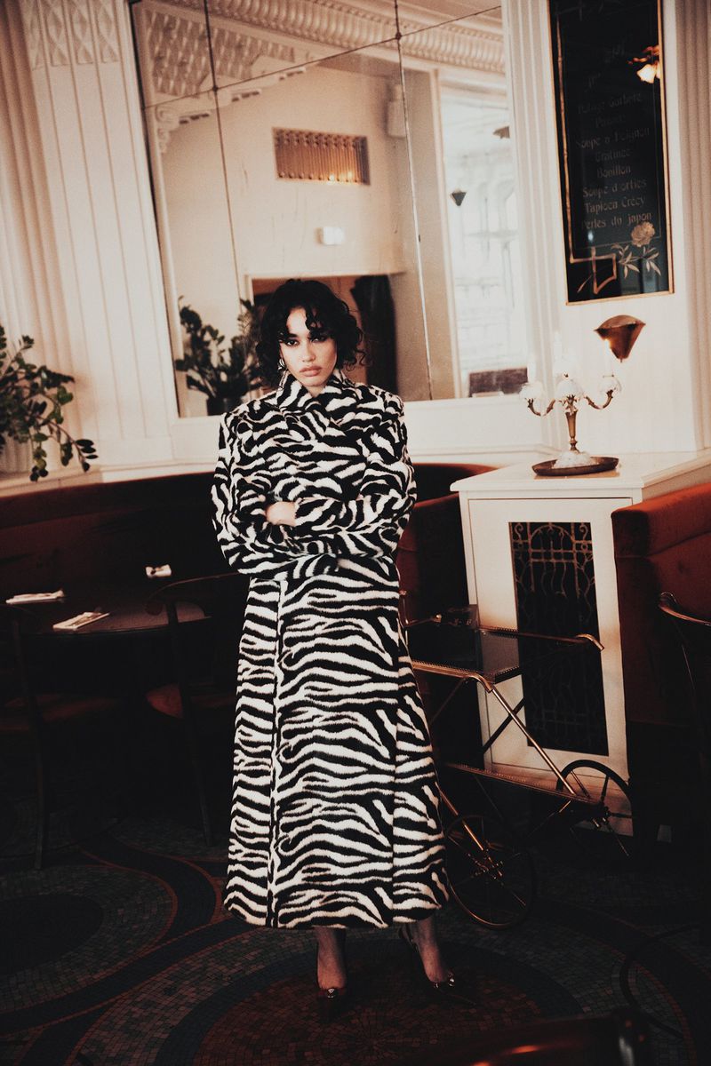 Adult female wearing a zebra-striped coat stands in an indoor environment, illuminated by natural light, complemented by a decorative table lamp.