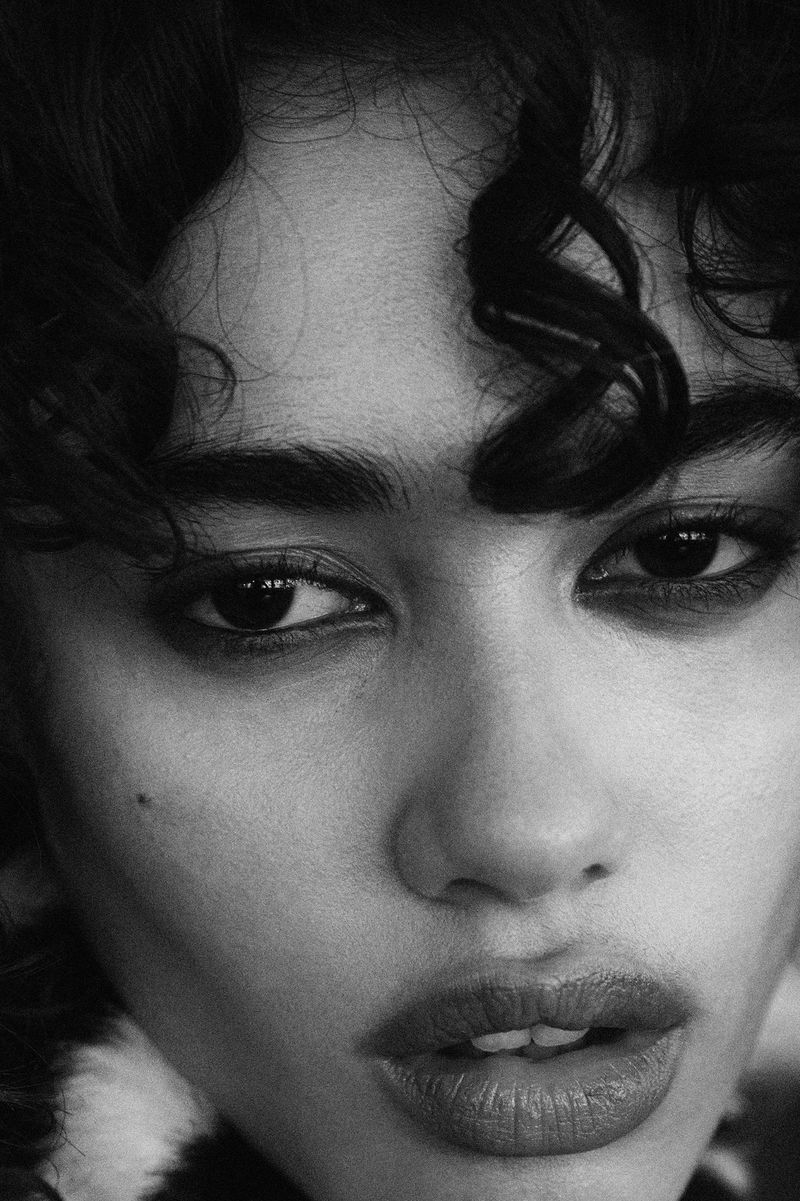 Striking black and white close-up portrait of a young woman with curly hair, dark eyes, and full lips in a beauty or fashion context.