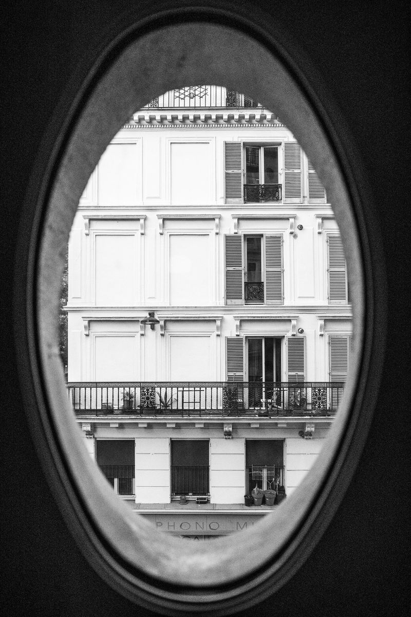 Black and white architectural shot from inside a building frames an exterior building with windows, shutters, and a small balcony.
