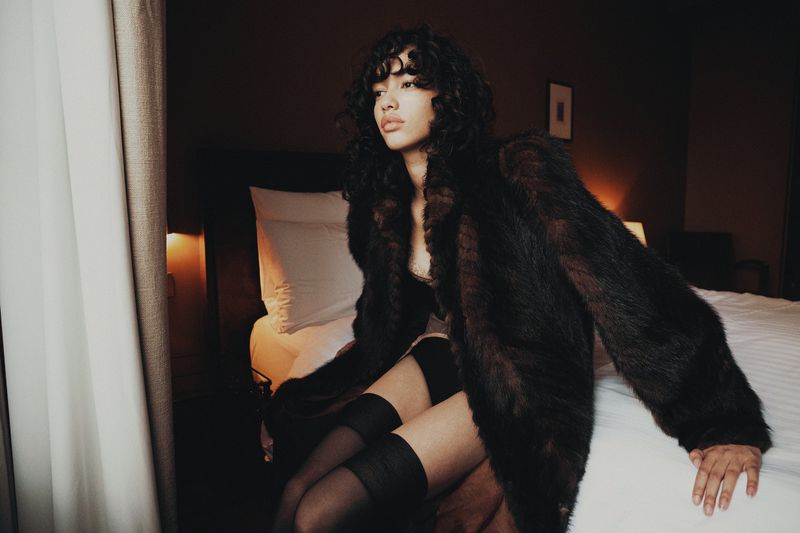 Elegant adult woman in a faux fur coat poses on a bed in a room with dark wood walls and sheer curtains, creating a moody, intimate scene.