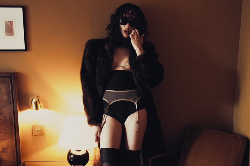 A woman poses indoors wearing lingerie, a fur coat, and sunglasses, with a lamp and framed art in the background, showcasing fashion.