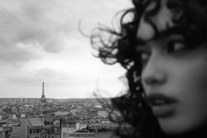 Black and white close-up profile of a woman with curly hair overlooking the Paris cityscape and Eiffel Tower in the distance.
