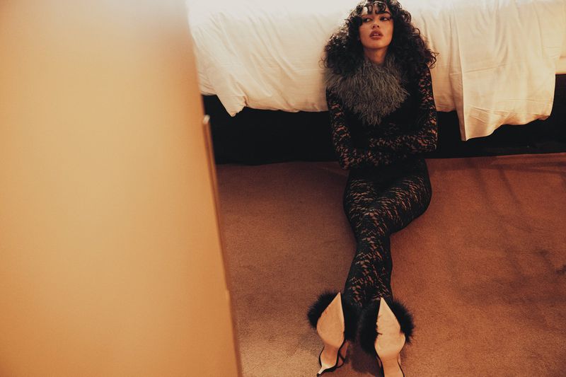 Elegant woman with curly hair in a black lace catsuit and heels poses on a brown carpet with a white bed in the background.