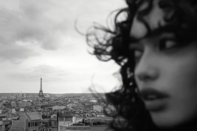Black and white close-up profile of a woman with curly hair overlooking the Paris cityscape and Eiffel Tower in the distance.
