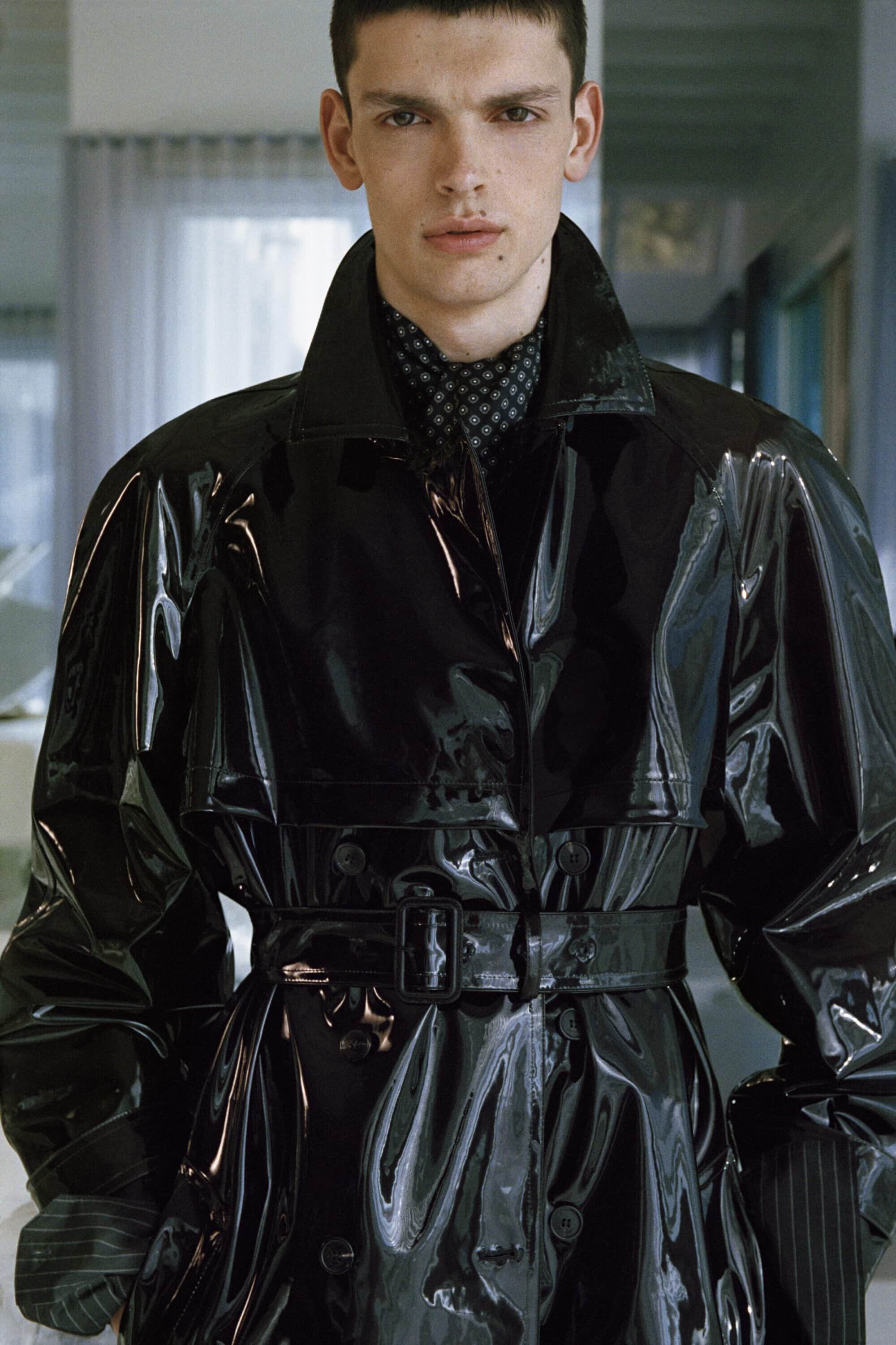 Model wearing a high-gloss black patent belted trench coat with a high collar and patterned scarf indoors.