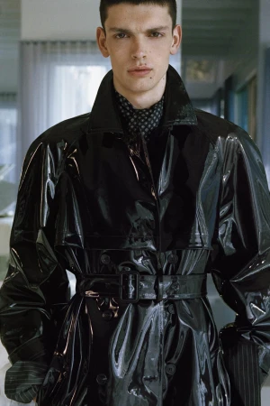 Model wearing a high-gloss black patent belted trench coat with a high collar and patterned scarf indoors.