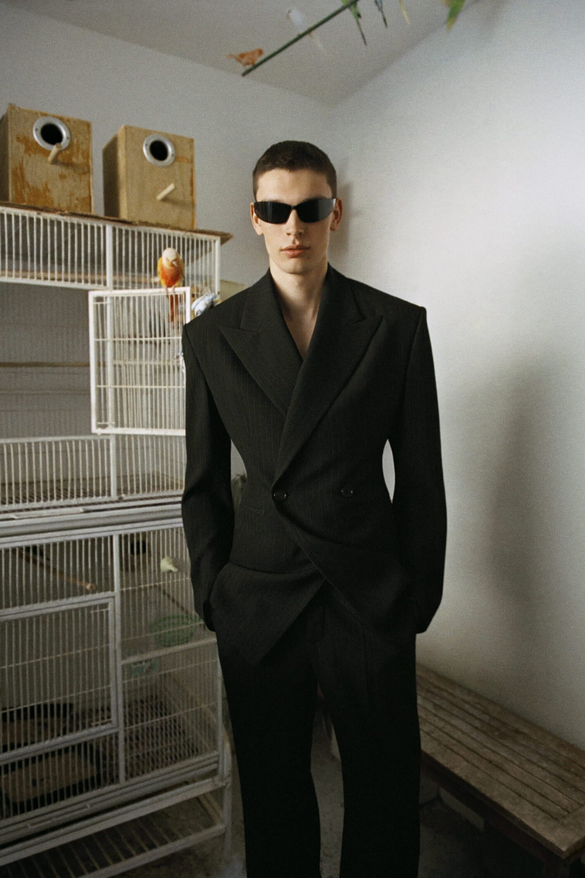 Man wearing dark sunglasses and a structured black suit standing indoors next to a large white birdcage.