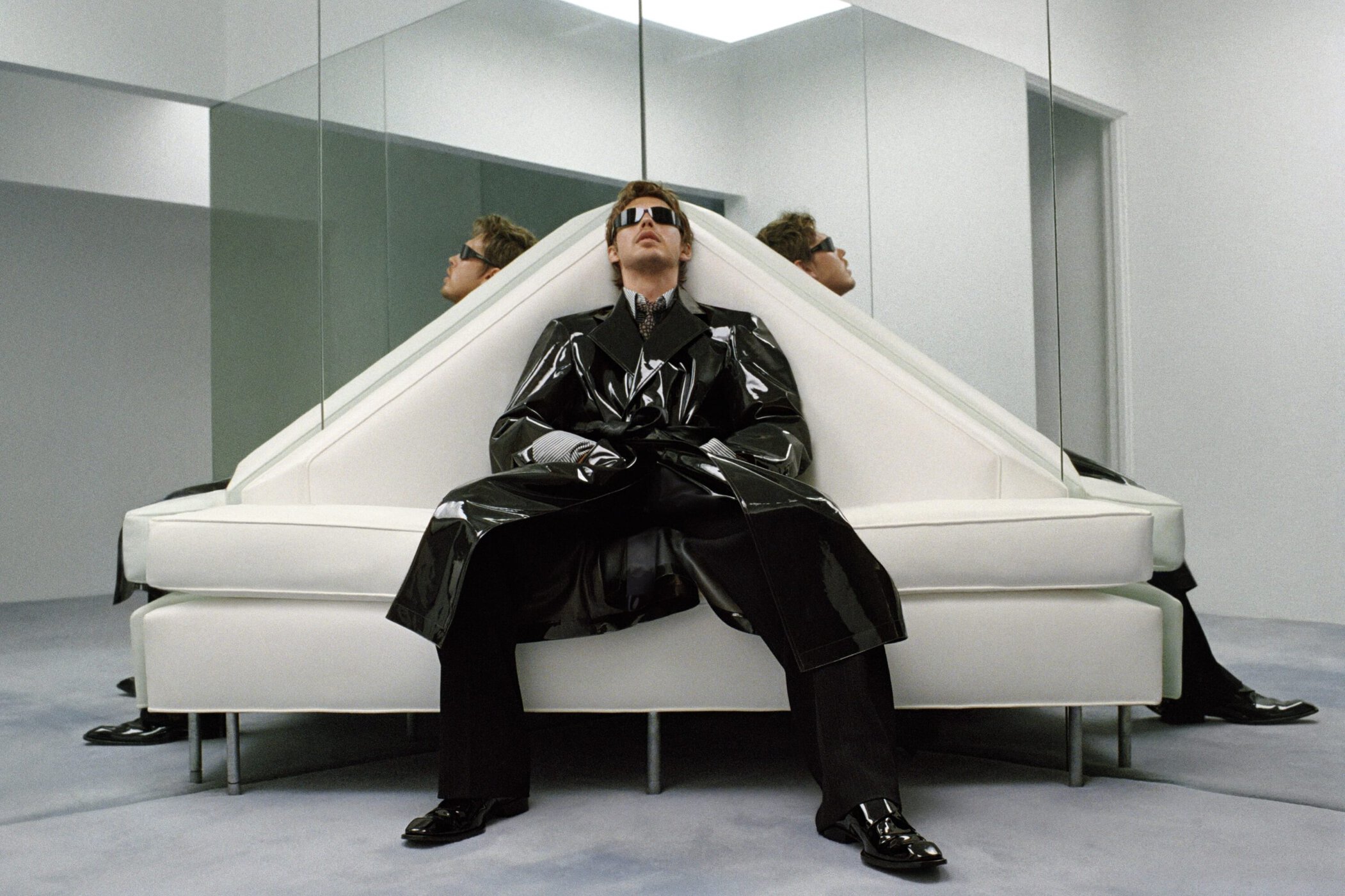 Austin Butler in a glossy black trench coat and sunglasses seated on a white geometric couch in a mirrored room.