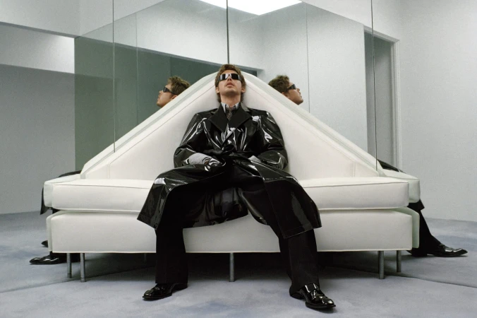 Austin Butler in a glossy black trench coat and sunglasses seated on a white geometric couch in a mirrored room.