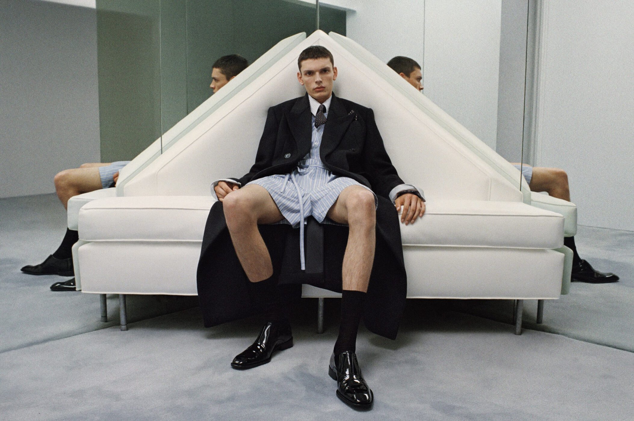 Model in a black overcoat and striped shorts seated on a white geometric sofa with mirror reflections.