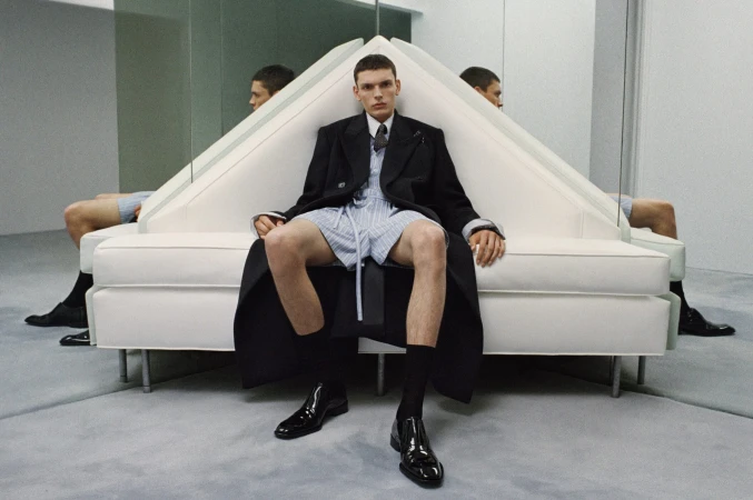 Model in a black overcoat and striped shorts seated on a white geometric sofa with mirror reflections.