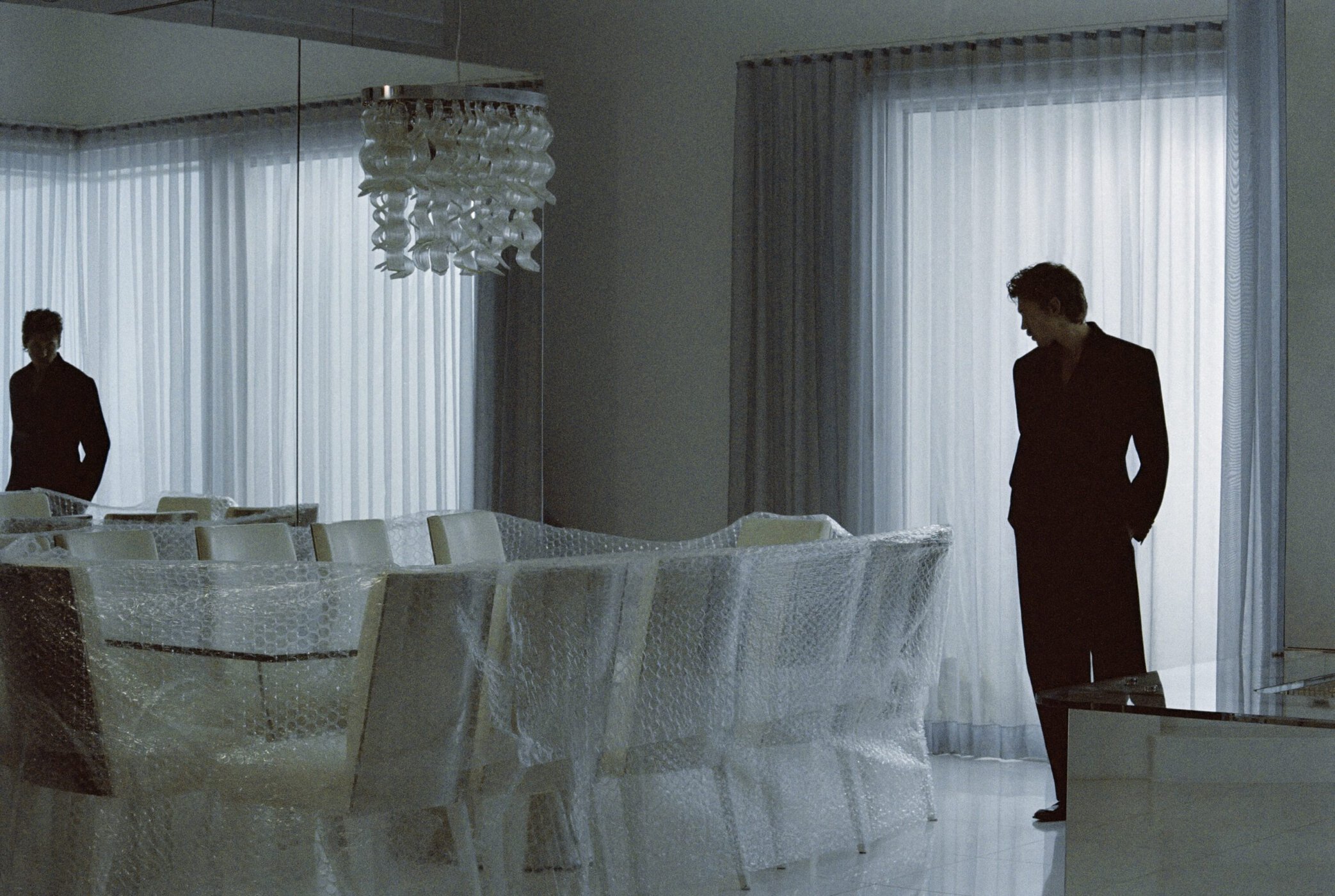 Austin Butler in a black suit standing in an interior next to bubble-wrapped dining chairs and sheer curtains.
