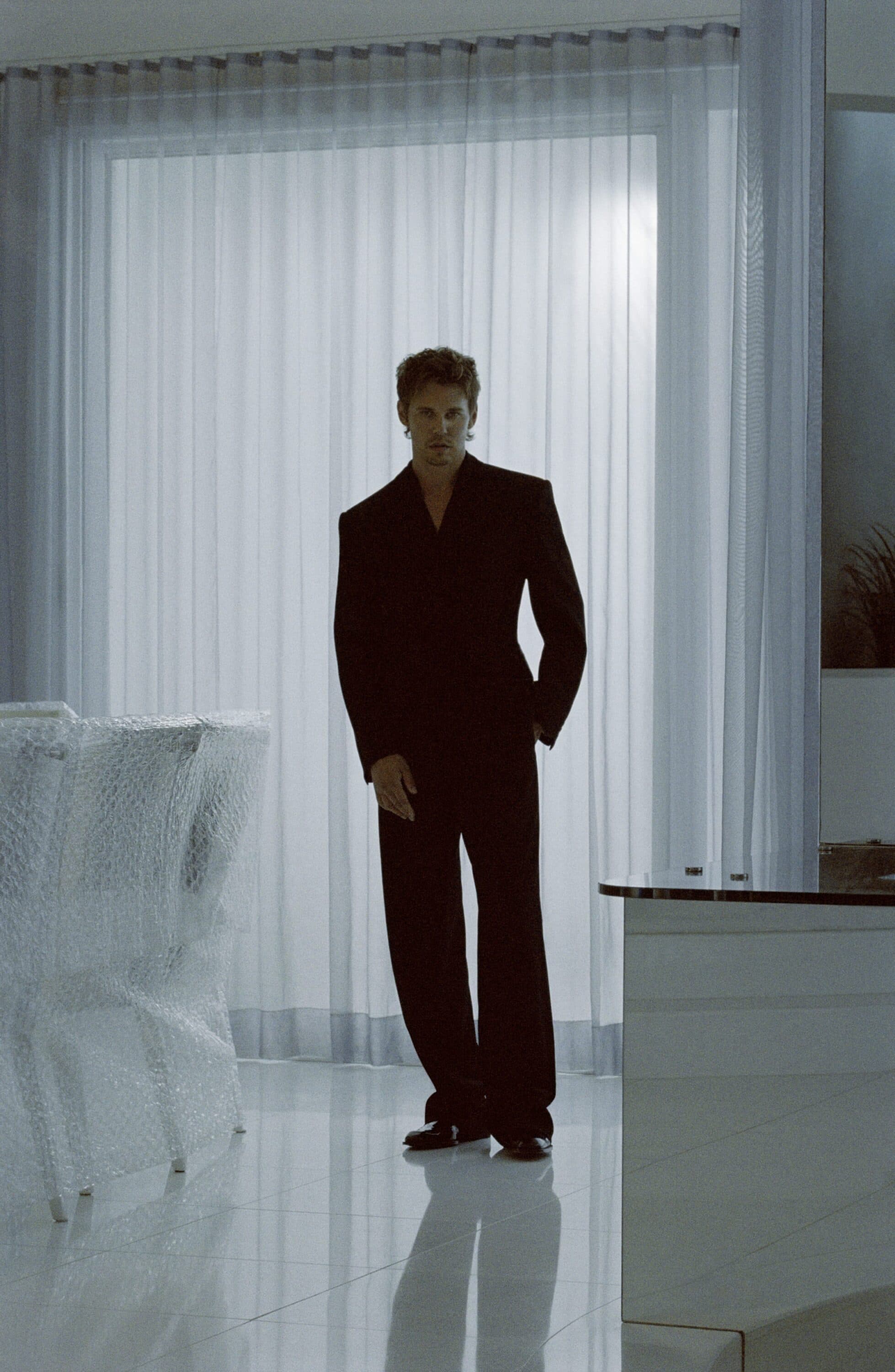 Austin Butler wearing a black tailored suit standing on a white tiled floor in front of sheer curtains.